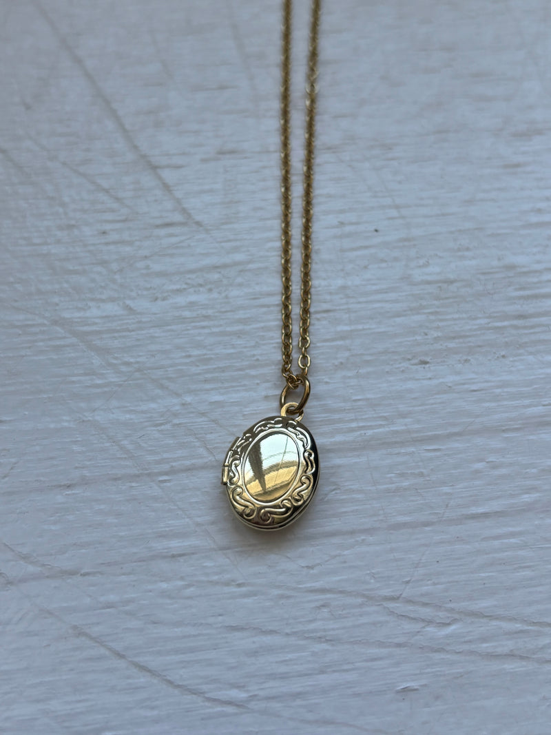 Gold-plated oval locket pendant necklace on an 18-inch stainless steel pvd coated gold chain, displayed on a white wooden surface. A vintage-inspired sentimental jewelry piece.