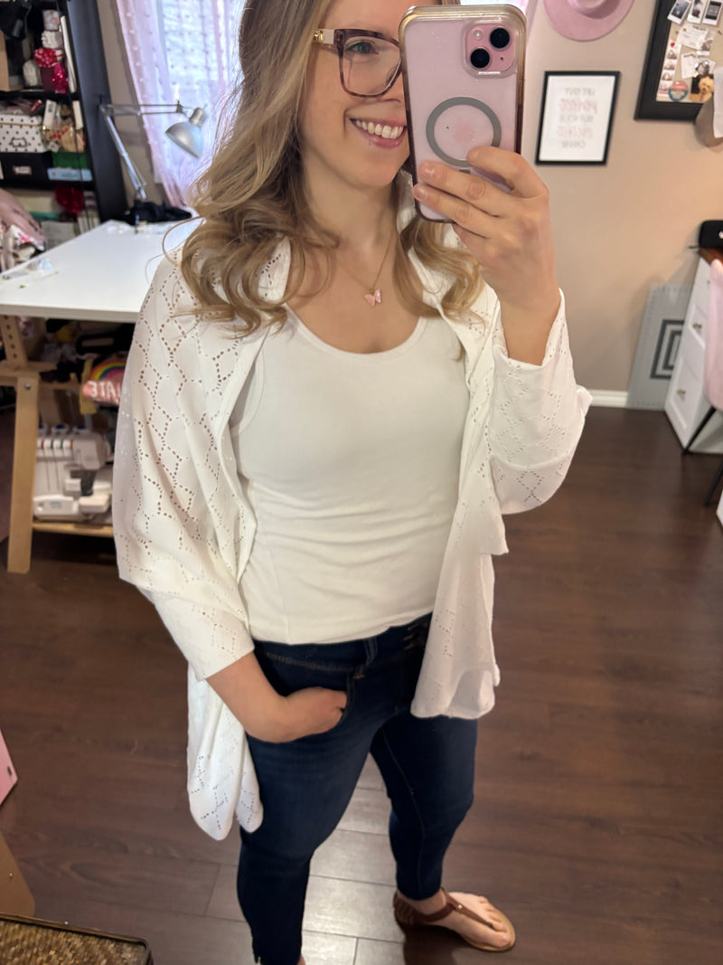 Smiling woman taking a mirror selfie while wearing a white City Shrug over a white tank top and dark jeans, paired with casual sandals.