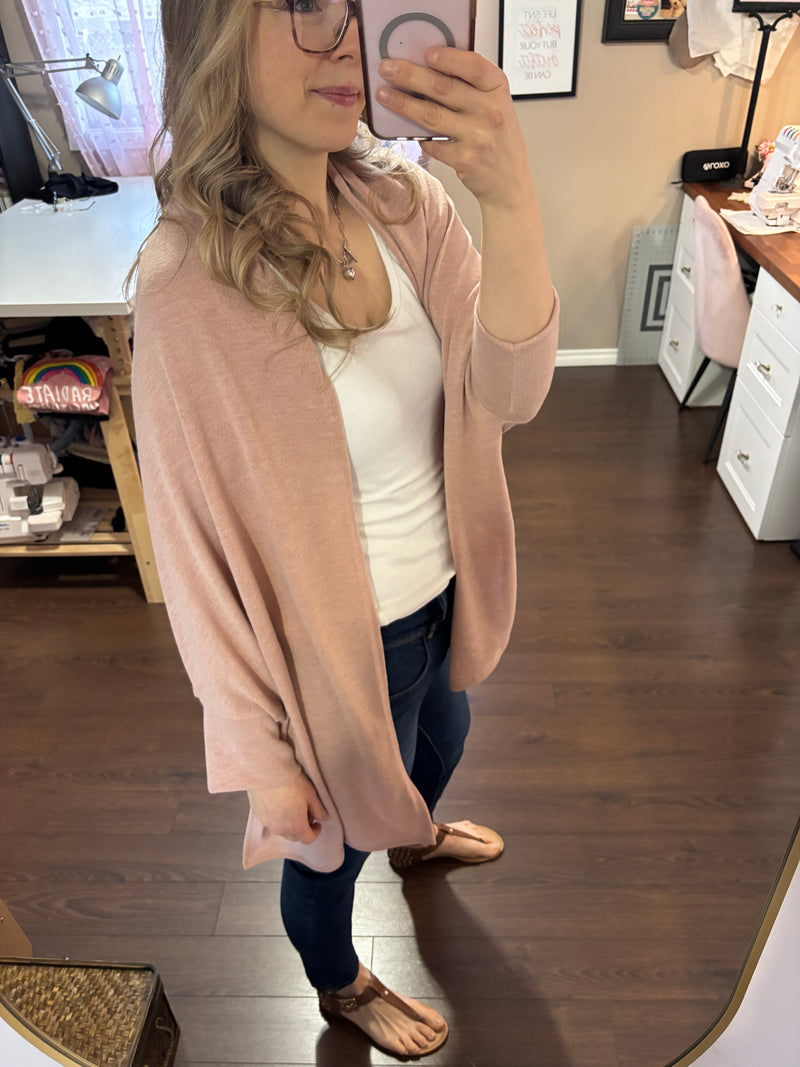 Smiling woman taking a mirror selfie while wearing a pink City Shrug over a white tank top and dark jeans, paired with casual sandals.