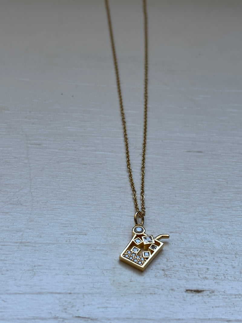 Gold-plated iced drink pendant necklace with crystal ice cube details on an 18-inch stainless steel pvd coated gold chain, displayed on a white wooden surface. A fun and trendy jewelry piece.