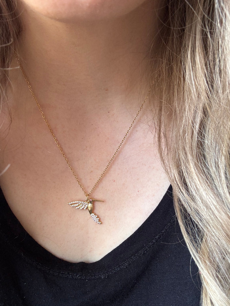 Gold-plated hummingbird pendant necklace with crystal details on an 18-inch stainless steel pvd coated gold chain, worn by a person. A delicate and symbolic nature-inspired jewelry piece