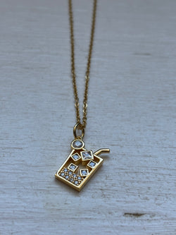 Gold-plated iced drink pendant necklace with crystal ice cube details on an 18-inch stainless steel pvd coated gold chain, displayed on a white wooden surface. A fun and trendy jewelry piece.