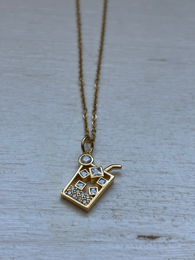 Gold-plated iced drink pendant necklace with crystal ice cube details on an 18-inch stainless steel pvd coated gold chain, displayed on a white wooden surface. A fun and trendy jewelry piece.