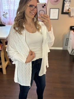 Smiling woman taking a mirror selfie while wearing a white City Shrug over a white tank top and dark jeans, paired with casual sandals.