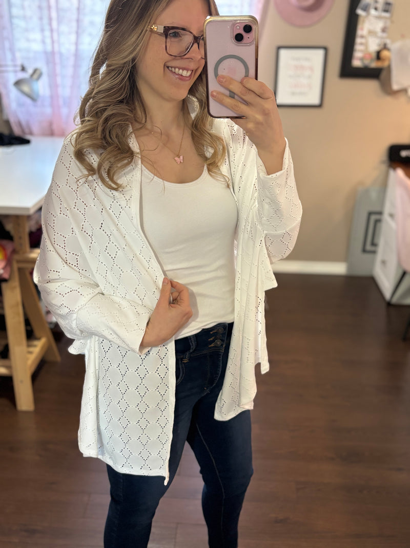 Smiling woman taking a mirror selfie while wearing a white City Shrug over a white tank top and dark jeans, paired with casual sandals.