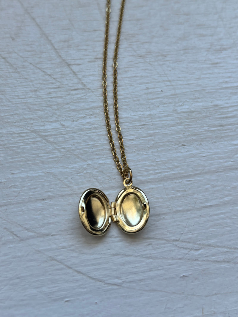 Gold-plated oval locket pendant necklace on an 18-inch stainless steel pvd coated gold chain, displayed on a white wooden surface. A vintage-inspired sentimental jewelry piece.
