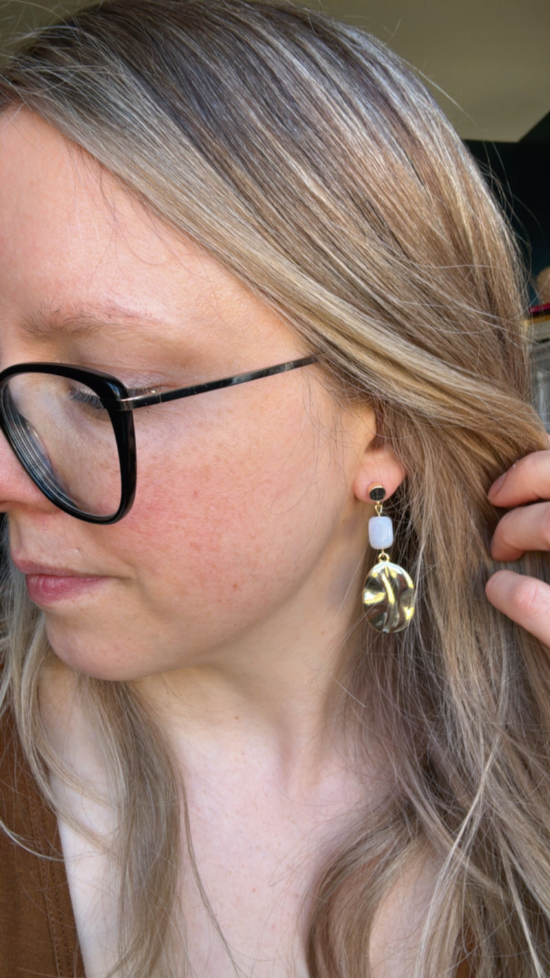 A close-up of a person with light blonde hair wearing the gold and agate dangle earrings. The earring features a rectangular gray agate stone with a hammered gold oval charm dangling below, adding a stylish and sophisticated touch to the look.