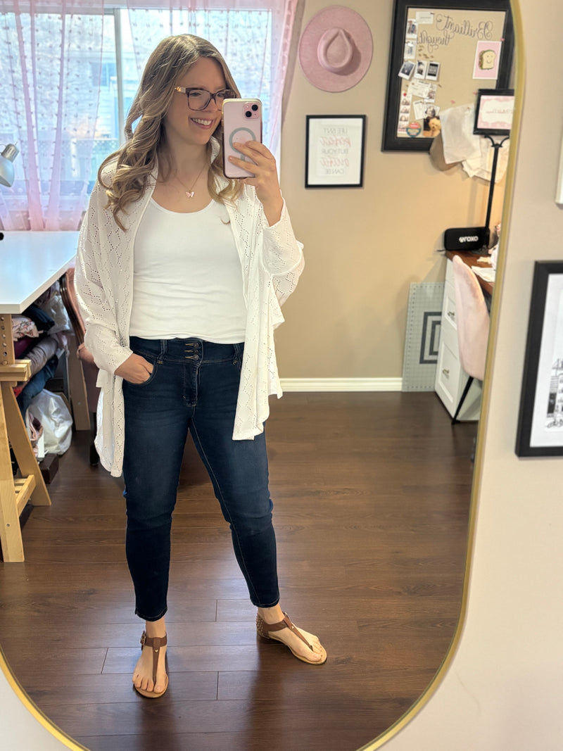 Smiling woman taking a mirror selfie while wearing a white City Shrug over a white tank top and dark jeans, paired with casual sandals.