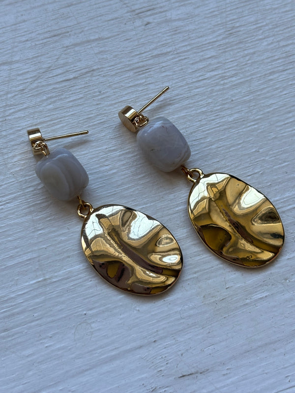  A pair of gold-plated dangle earrings featuring a rectangular gray agate stone at the top and a hammered gold oval charm hanging below. The earrings are displayed on a white wooden surface, showcasing their elegant and modern design.