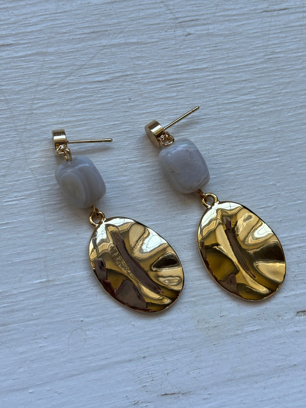  A pair of gold-plated dangle earrings featuring a rectangular gray agate stone at the top and a hammered gold oval charm hanging below. The earrings are displayed on a white wooden surface, showcasing their elegant and modern design.