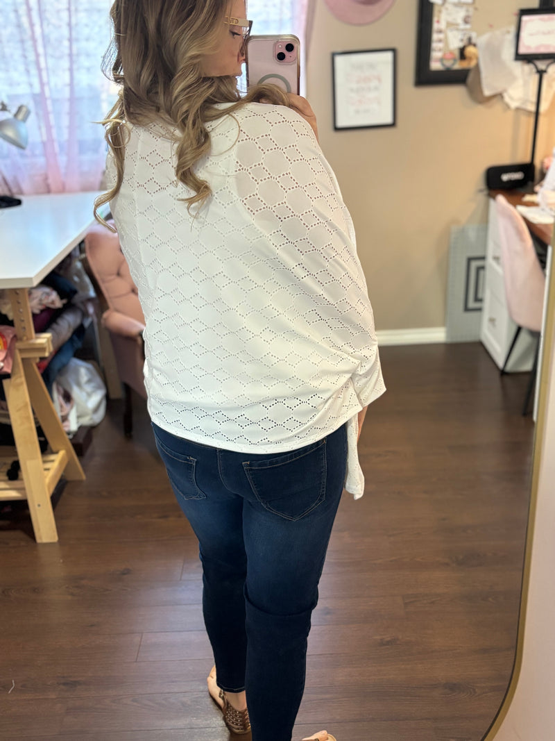 Woman wearing a white eyelet-patterned City Shrug with a draped back, styled with dark jeans in a cozy, feminine workspace.